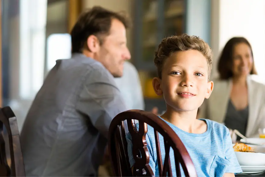 Junge sitzt auf einem Stuhl, im Hintergrund sind seinem Eltern am Tisch