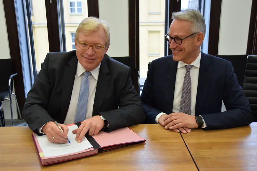 Dr. Ralf Kleindiek und Prof. Dr. Harald Schmitz sitzen an einem Tisch, Dr. Ralf Kleindiek unterzeichnet einen Vetrag
