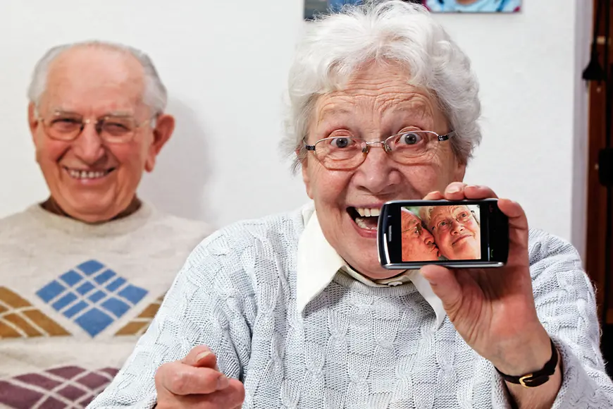 Ein älteres Liebespaar. Die Frau hält ein Smartphone in die Kamera, auf dem ein Foto mit dem küssenden Paar zu erkennen ist.