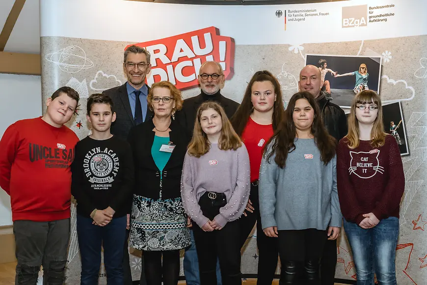 Das Bild zeigt mehrere Erwachsene und Kinder vor einem Plakat der Initiative "Trau dich!"