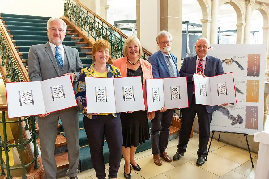 Das Bild zeigt Caren Marks und vier Personen, die vor einer Treppe stehend die Briefmarkenerstdrucke in den Händen halten