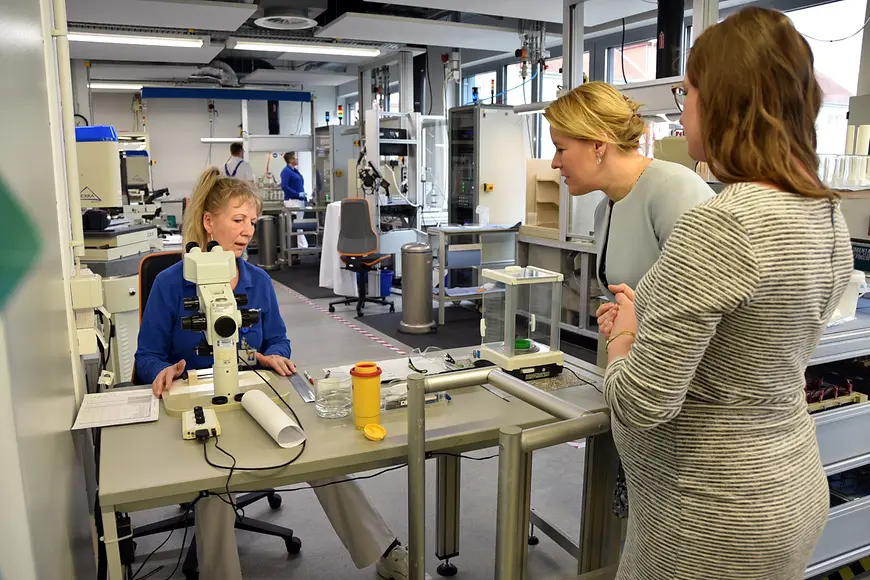 Eine Mitarbeiterin von Cortronik sitzt an ihrem Arbeitstisch und erklärt ihre Arbeit. Dr. Franziska Giffey hört ihr zu.