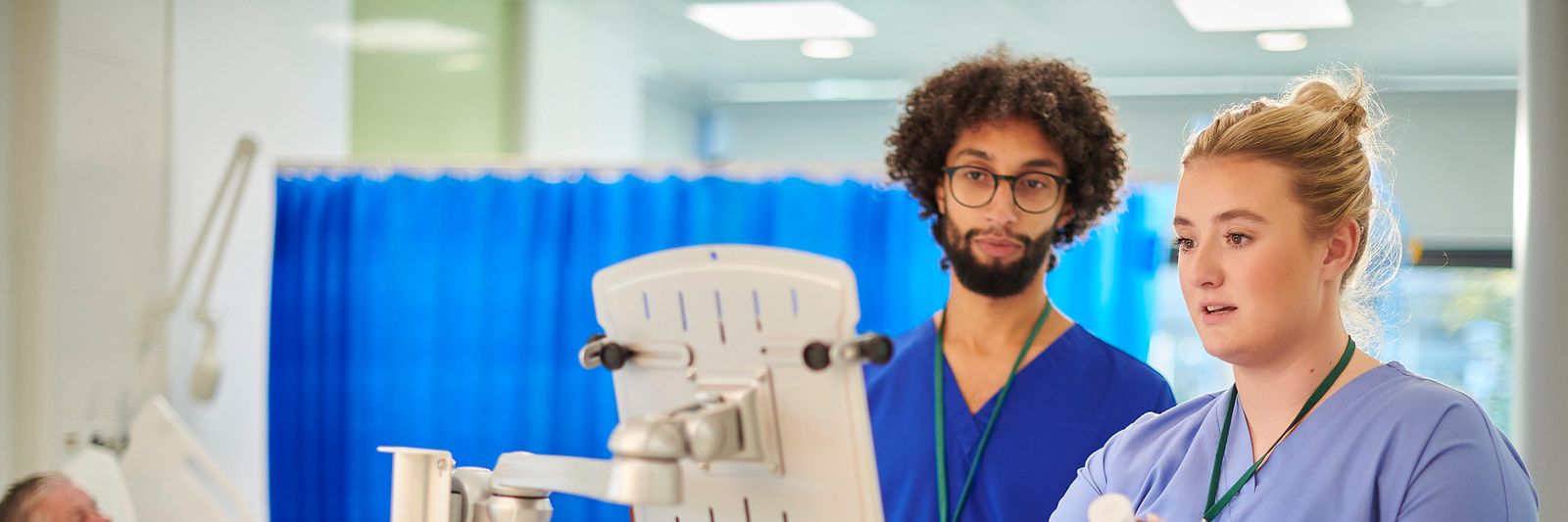 Zwei pflegende Personen und ein Mensch in einem Krankenbett im Hintergrund