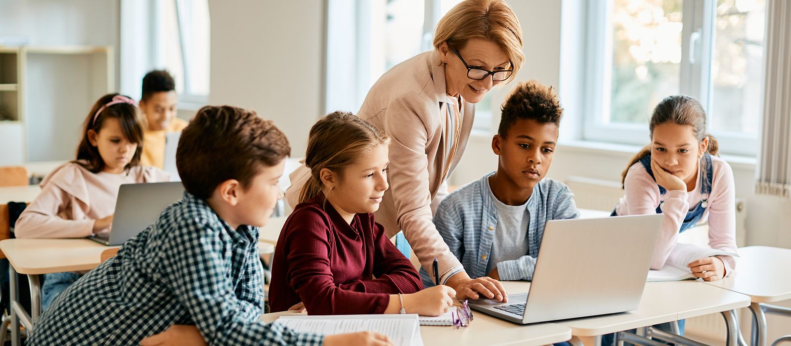 Mehrer Schulkinder schauen auf den Bildschirm eines Laptops, während ihnen die Lehrerin dazu etwas erklärt.