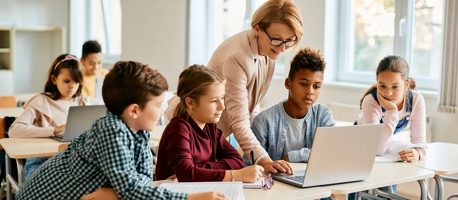 Mehrer Schulkinder schauen auf den Bildschirm eines Laptops, während ihnen die Lehrerin dazu etwas erklärt.