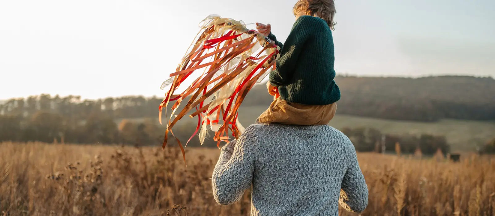 Ein Kind sitzt auf den Schultern eines Erwachsenen. Das Kind hält einen gebastelten Stock mit bunten Bändern in der Hand.
