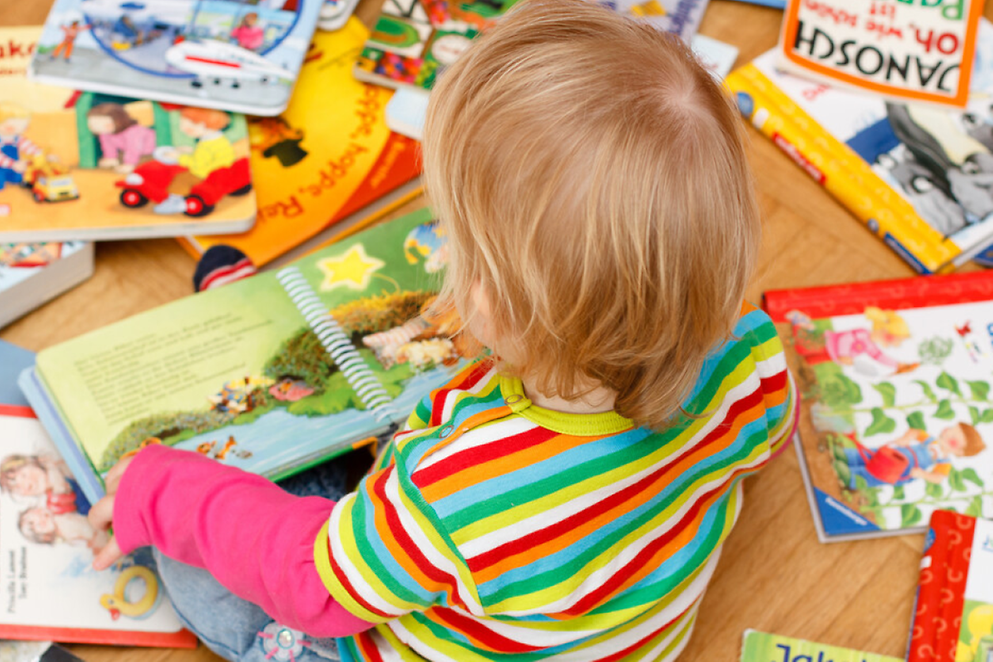 Rückansicht eines kleinen Kindes, das auf dem Boden sitzt und ein Kinderbuch aufgeschlagen hat
