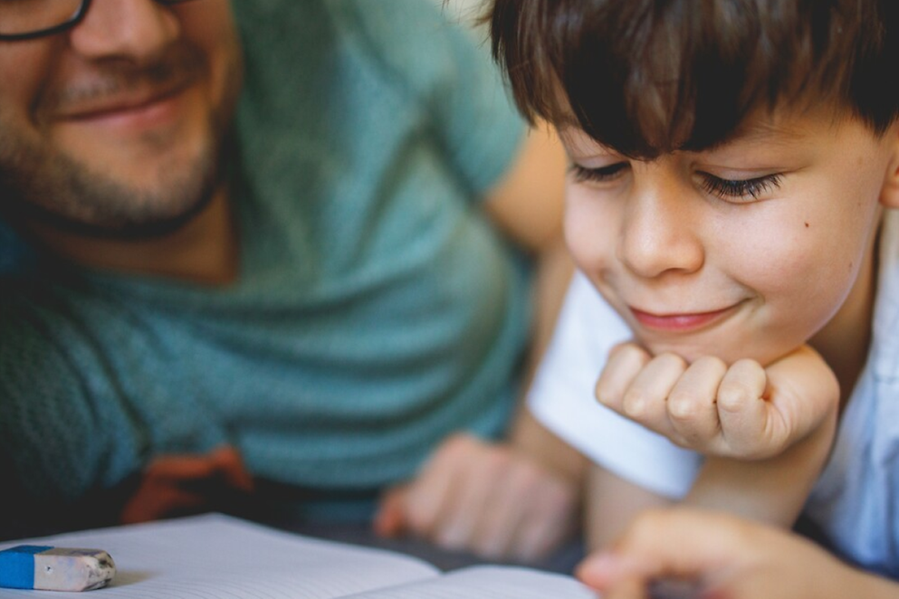 Ein Kind schaut lächelnd in ein Buch und stützt den Kopf auf die Hand. Der Vater lächelt.