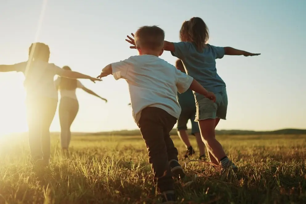 Fünf Kinder rennen mit ausgebreiteten Armen über eine Wiese dem Sonnenlicht entgegen