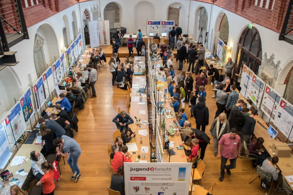 Luftaufnahme einer Veranstaltung mit Menschen an Tischen und Ständen in einem großen Raum