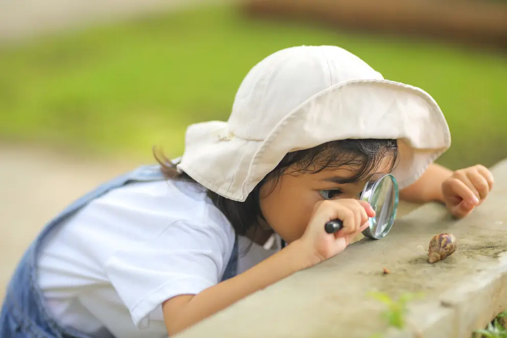 Ein junges Mädchen mit Hut untersucht mit einer Lupe eine Schnecke