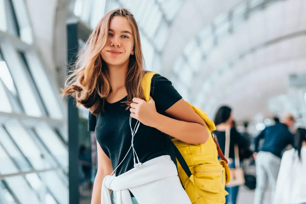 Eine junge Frau mit blondem Haar trägt ein T-Shirt und einen gelben Rucksack. Sie lächelt in die Kamera.