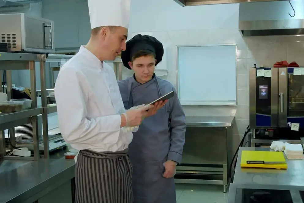 Ein Koch erklärt in einer Gastro-Küche einem Azubi etwas auf einem Tablet.