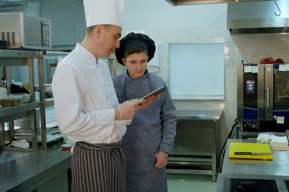 Ein Koch erklärt in einer Gastro-Küche einem Azubi etwas auf einem Tablet.