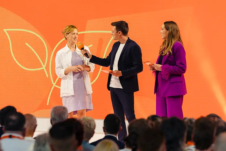 Karin Prien mit Moderator vor orangem Hintergrund
