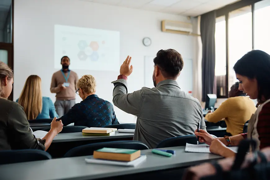 Ein Lehrer erklärt etwas vor einer Gruppe Erwachsener in einem Klassenraum.