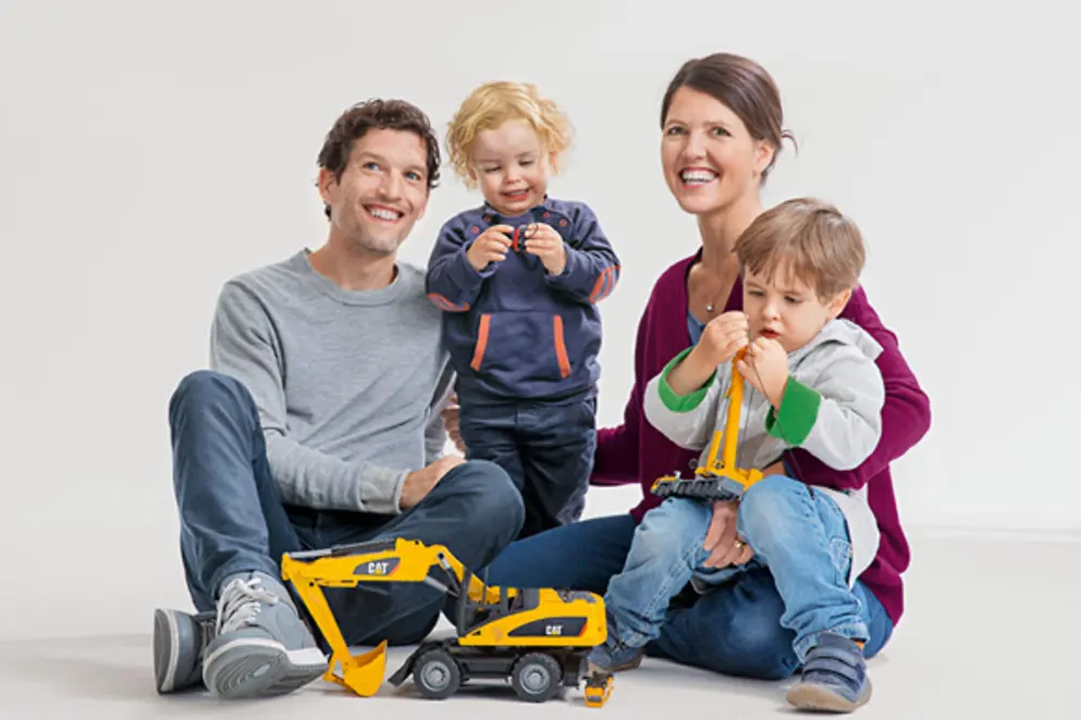 Eine junge Familie sitzt zusammen. Vater, Mutter und die beiden Kinder strahlen und spielen mit Legosteinen.