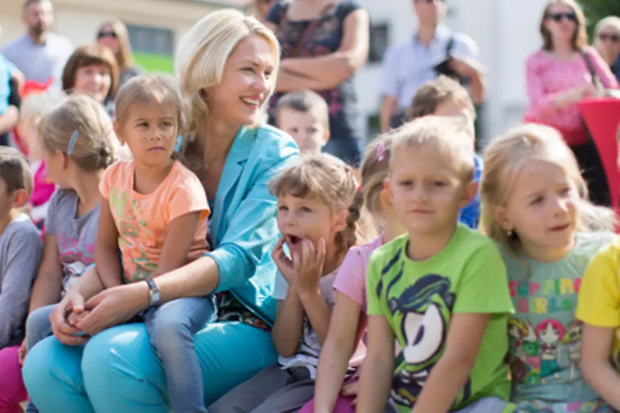 Alt: Gruppenbild mit Manuela Schweisg und Kindern in einer Kita. Ein junges Mädchen sitzt auf dem Schoß der Ministerin.